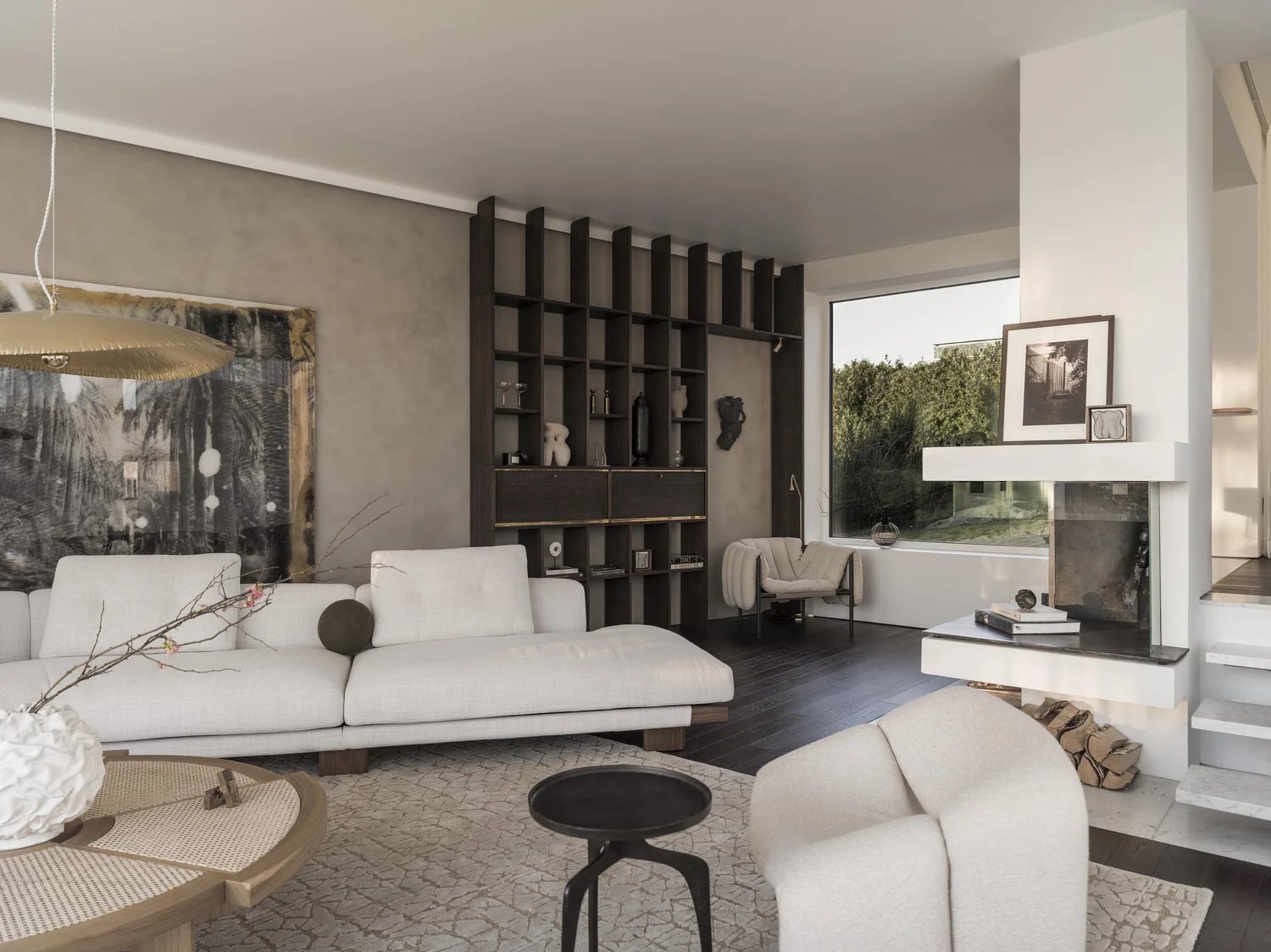 Modern living room with white sofa, beige armchair, dark wood shelving, marble fireplace, large window, and decorative items.