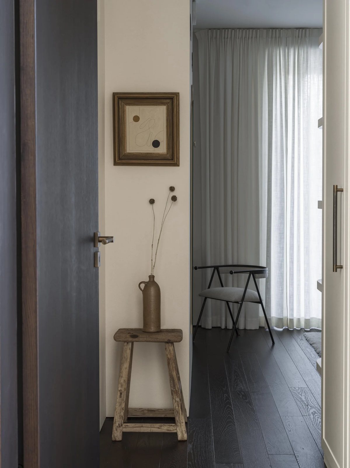 Rustic wooden stool in a softly lit hallway with framed art and natural light from sheer curtains.