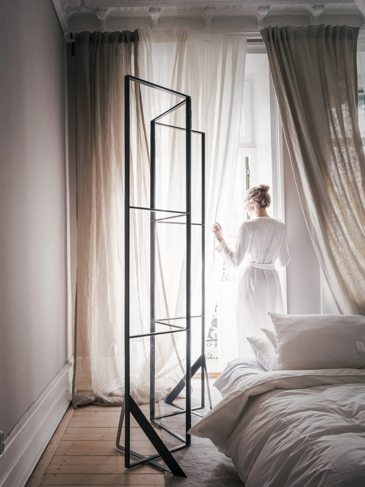 Woman in robe drawing linen curtains in a softly lit room with clean lines.