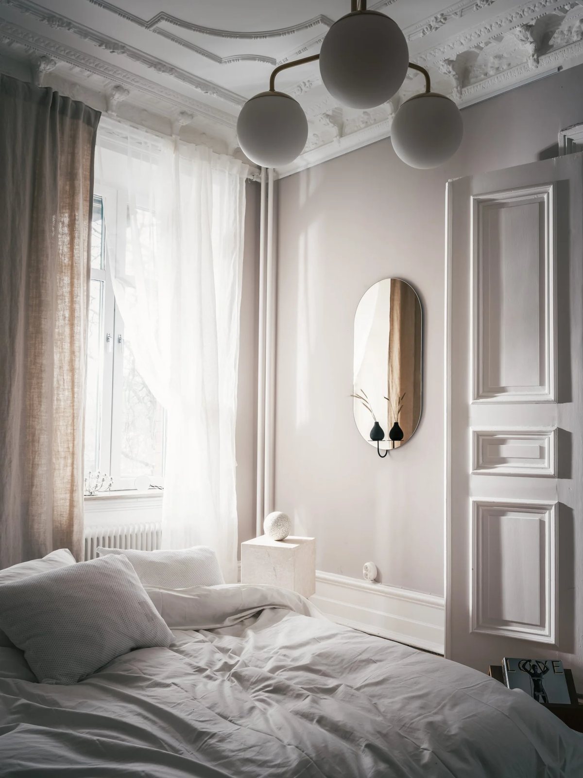 Detail of bedroom corner with wall mirror, sculptural lamp and linen curtains.