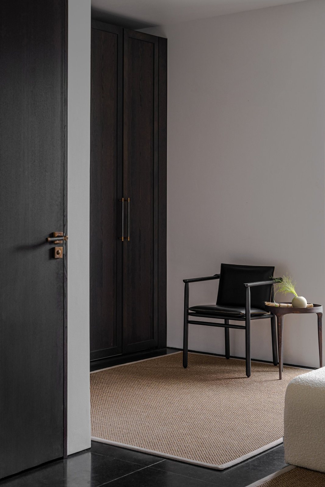 Minimalist corner with a dark wood wardrobe, black leather chair, and neutral rug.