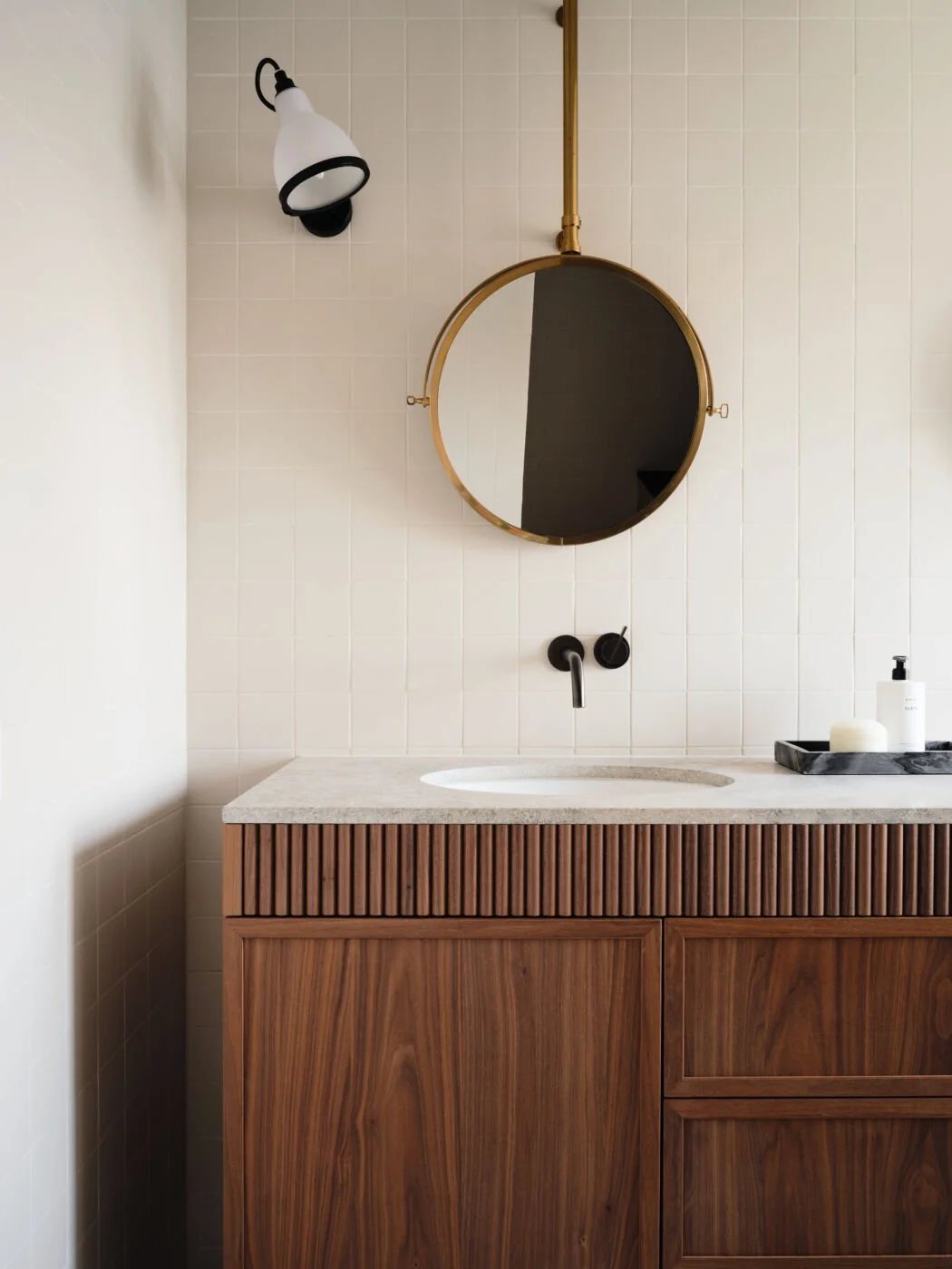 A minimal guest bathroom with fluted walnut cabinetry, creamy tiles, and a brass-framed round mirror suspended from the ceiling.