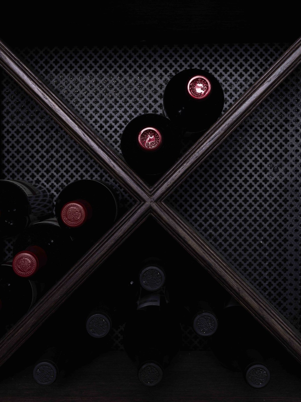 A geometric wine rack with X-shaped compartments holding dark wine bottles against a patterned mesh backdrop.