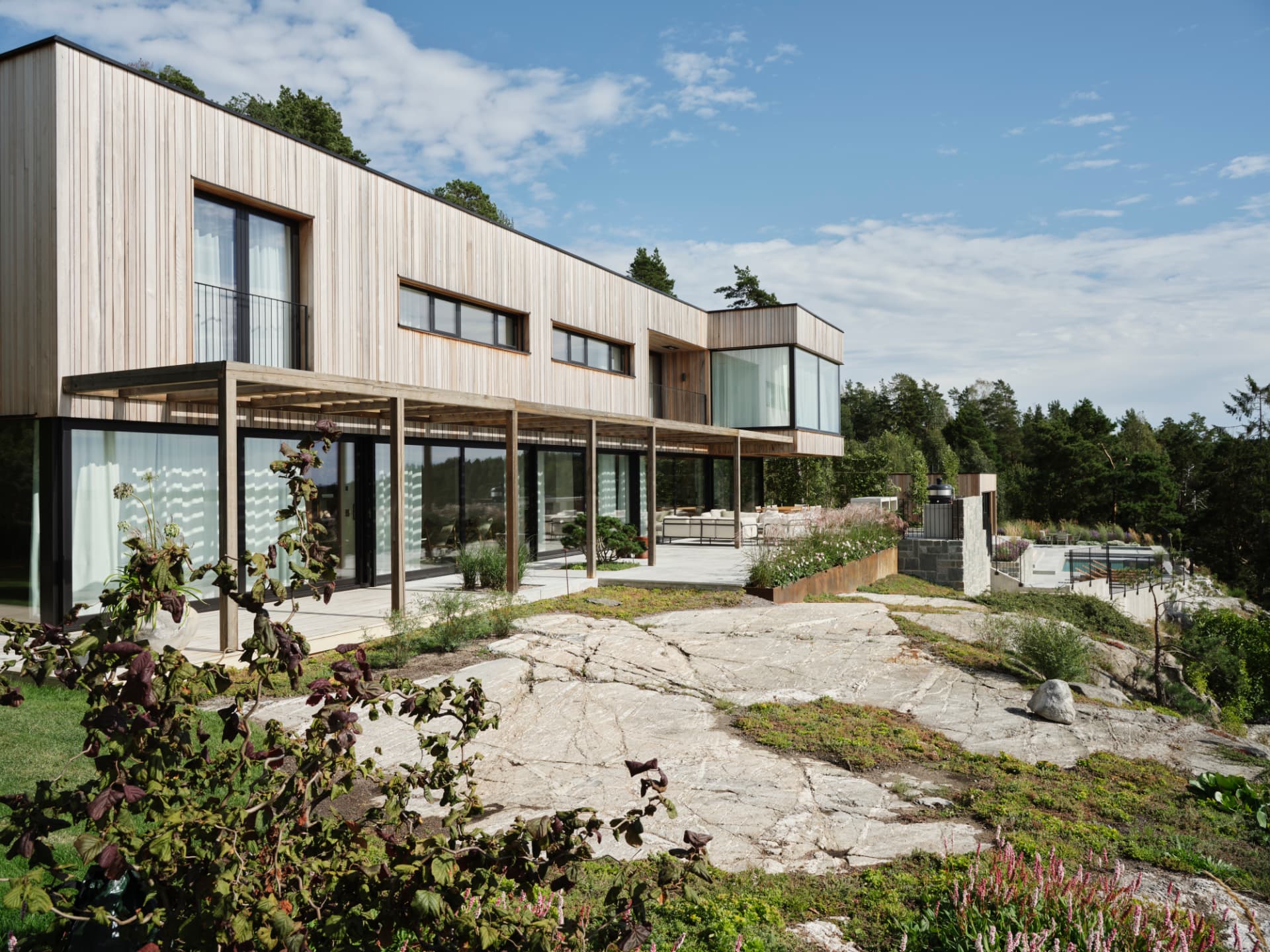 Timber-clad villa standing on exposed bedrock, with a pergola of pale posts, floor-to-ceiling glass and a pool beyond low stone walls