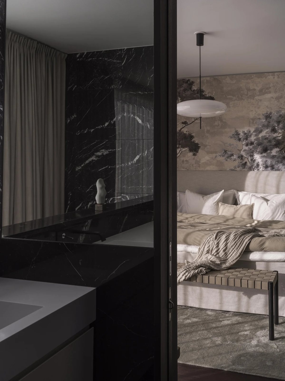 View from a black marble bathroom into a serene bedroom featuring a mural wall and linen bedding.