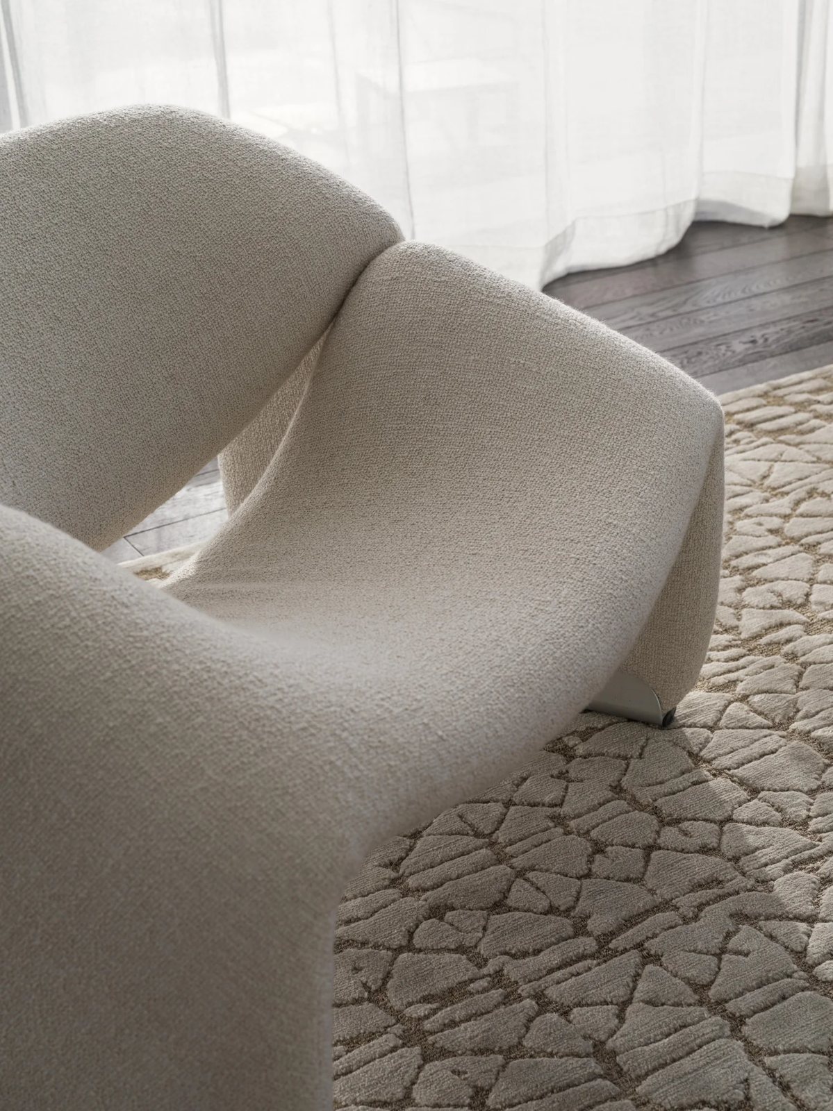 Close-up of a soft sculptural chair on a textured rug near sheer curtains.