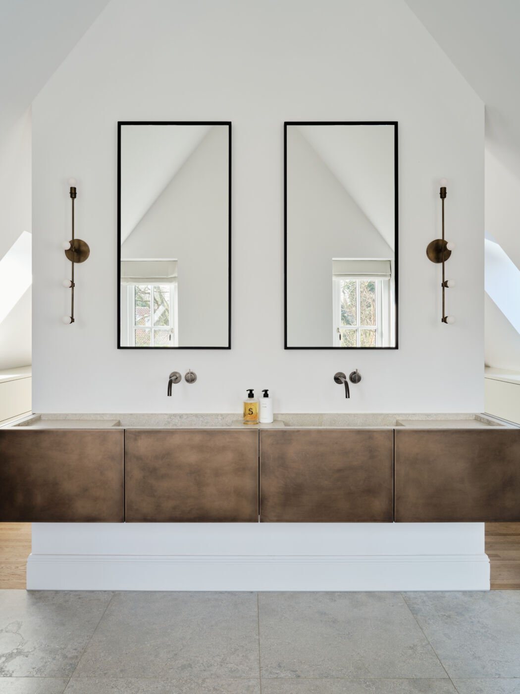 A sleek bathroom interior showcasing twin mirrors, wall-mounted taps and floating bronze cabinetry beneath a pitched ceiling.